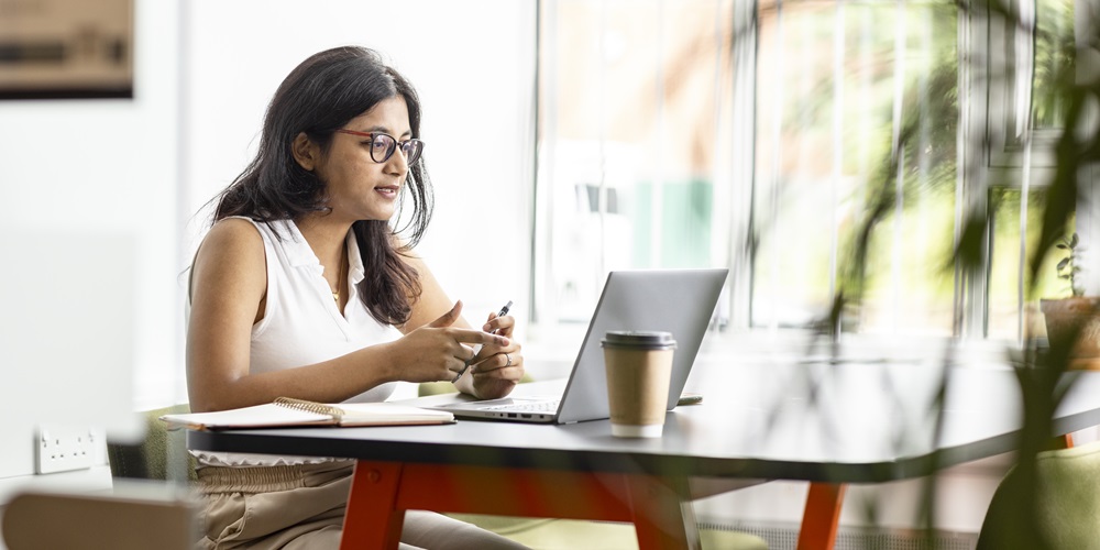 A student sat at a modern desk with a cup of coffee. They are on a video call via their laptop.