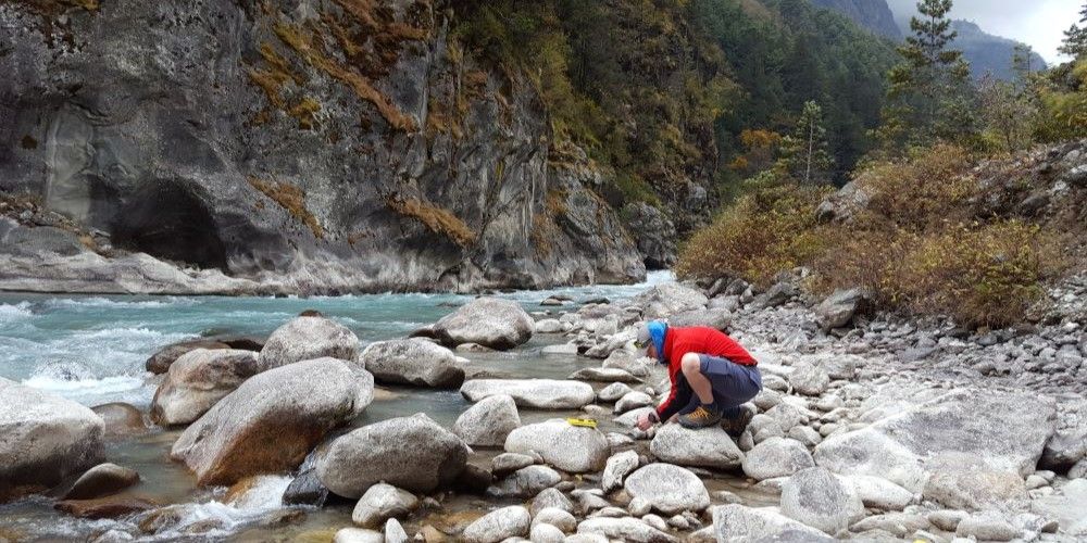 A researcher kneels on rocks next to a river in a valley, studying the river and rocks below.