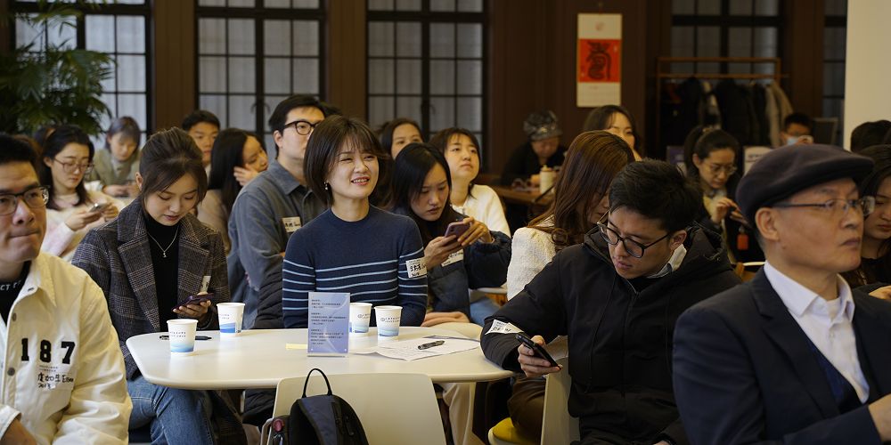 A group of people sat at round tables in a conference room watch a speaker at the front of the room