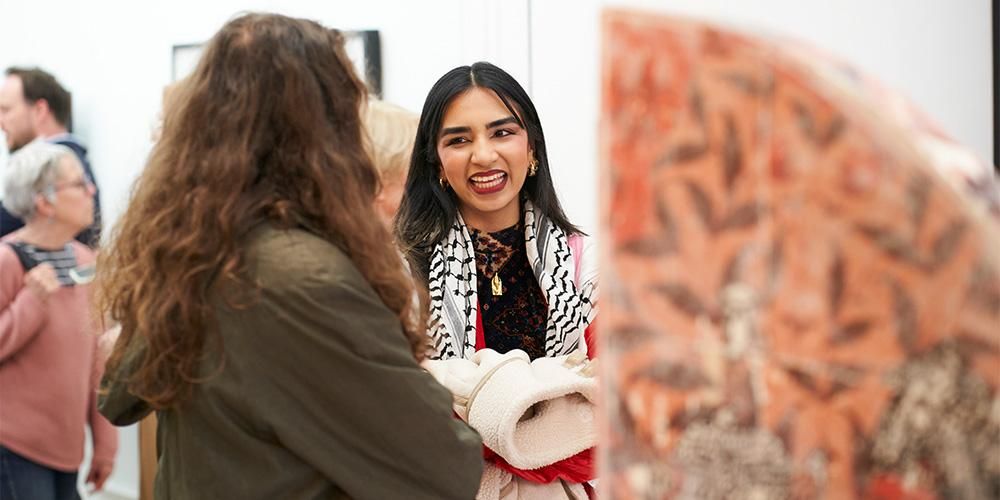 Two people smile at each other in conversation while they look at artwork on display at a gallery.