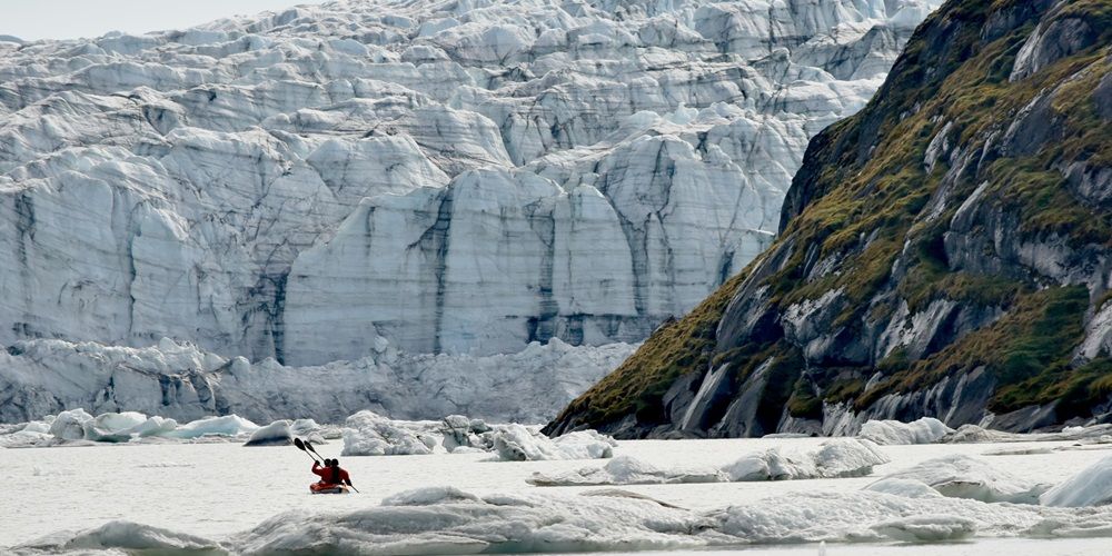 A glacial landscape with huge dramatic cliffs of rock and ice. Two people are rowing a small kayak in the foreground.