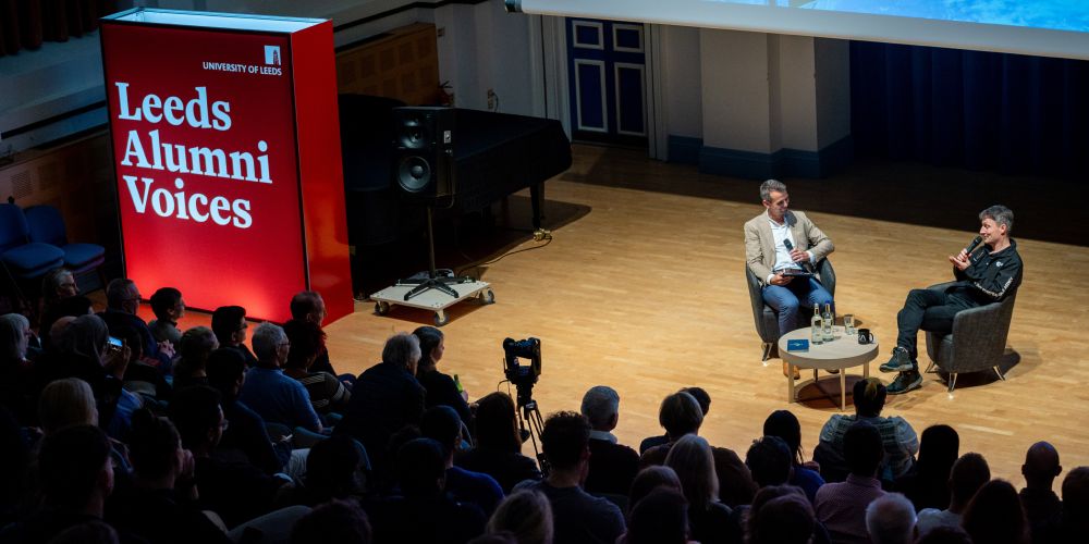 Two people sit on stage holding microphones and talking, with audience watching on. A sign on the stage reads 'Leeds Alumni Voices'.