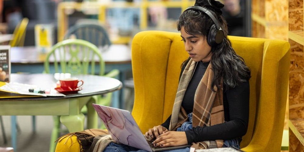 A student sat in a big comfy chair in cafe, wearing headphones and using a laptop.