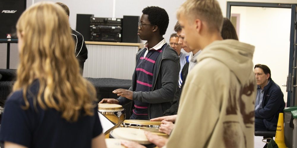 Visitors at an open day in a room filled with music equipment. Some are playing hand drums.