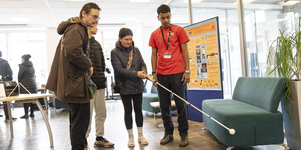 A group of people at open day. One person is using a white cane to explore a room.