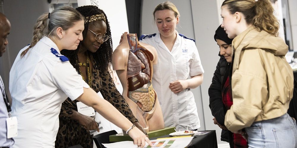 A group of people at open day gathered around a large 3D human anatomical model. Some are medical or healthcare students