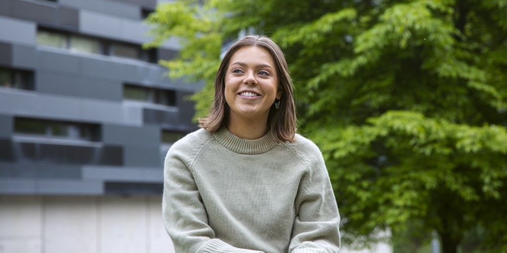 A smiling student sat down outside on campus with a modern building and tree behind them.