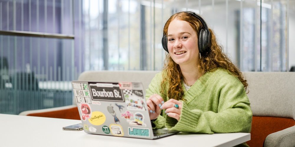 A student sat at a desk in a study space using a laptop and headphones and smiling