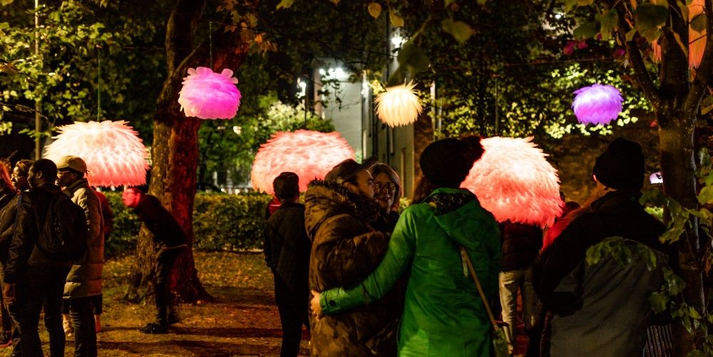 Multisensory, colourful, illuminated artwork is hung from trees with visitors looking at the art.