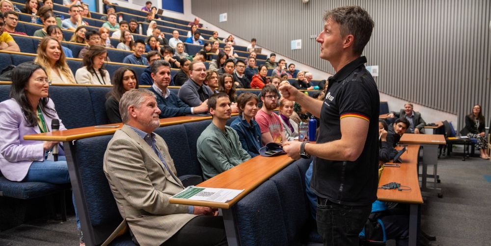 A person standing at the front of a lecture theatre, gesticulating and talking to people sat in rows of tiered seating.