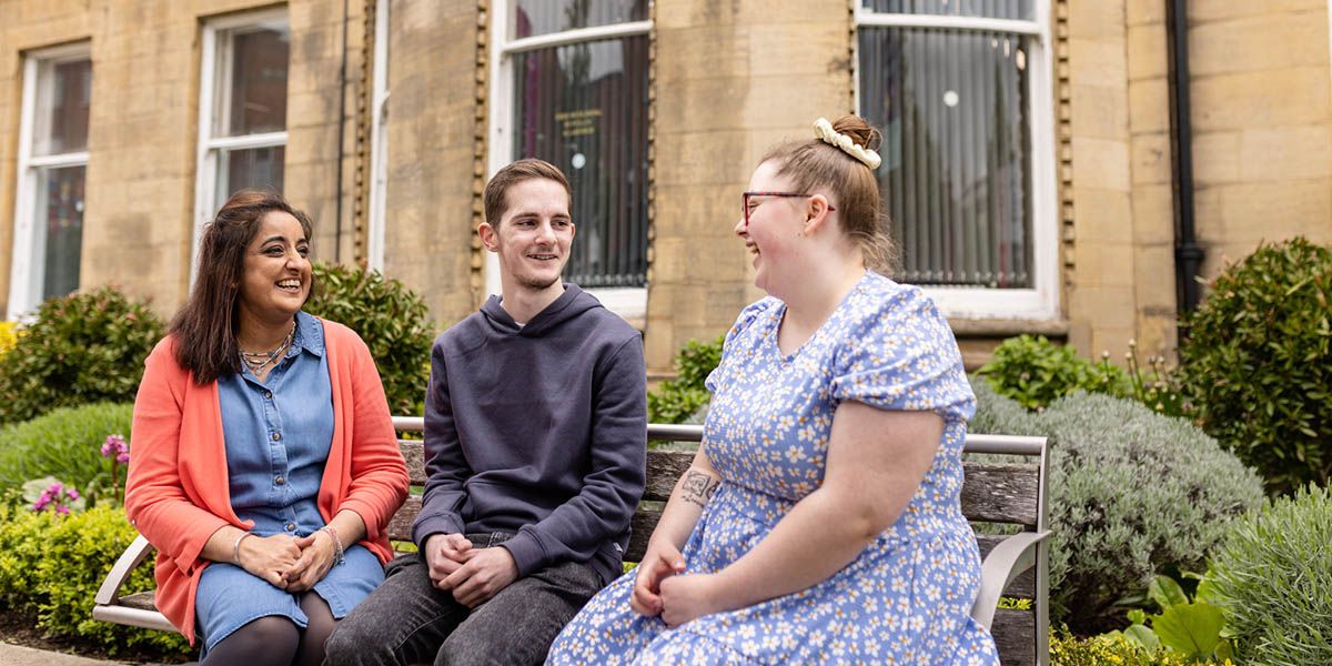 Three students smiling while sat on a bench on campus.