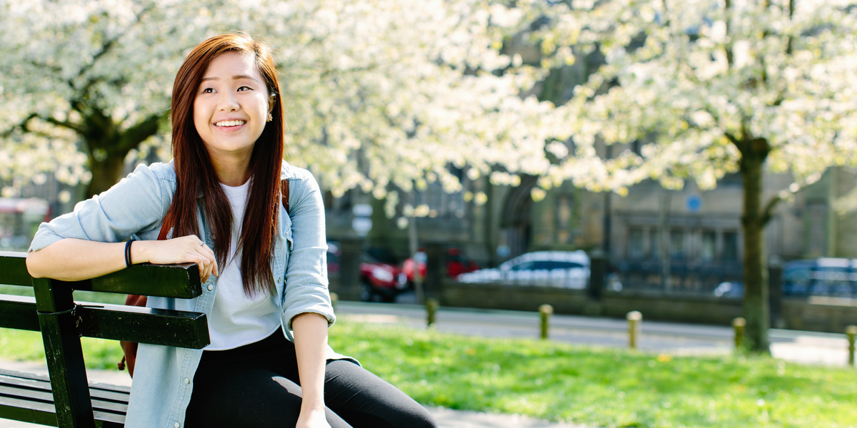 一名学生微笑着坐在樱花树旁的长椅上。