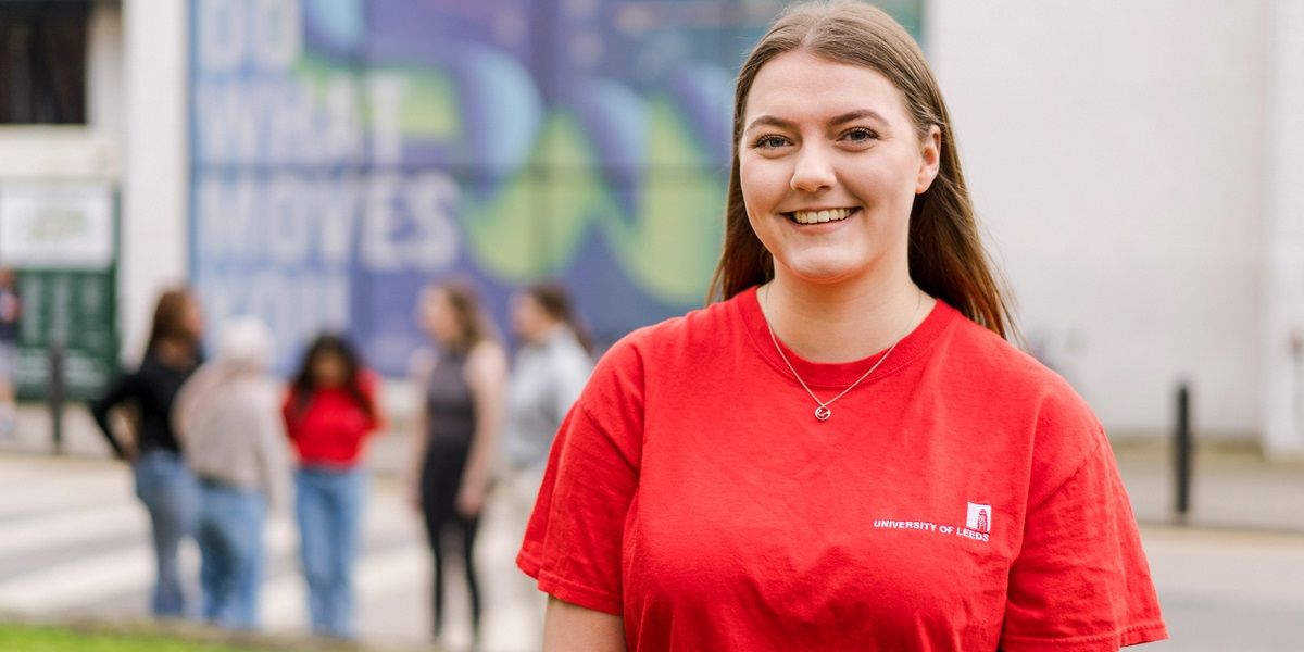 A student ambassador in red t-shirt stood on campus on open day. Open day visitors are in the background.