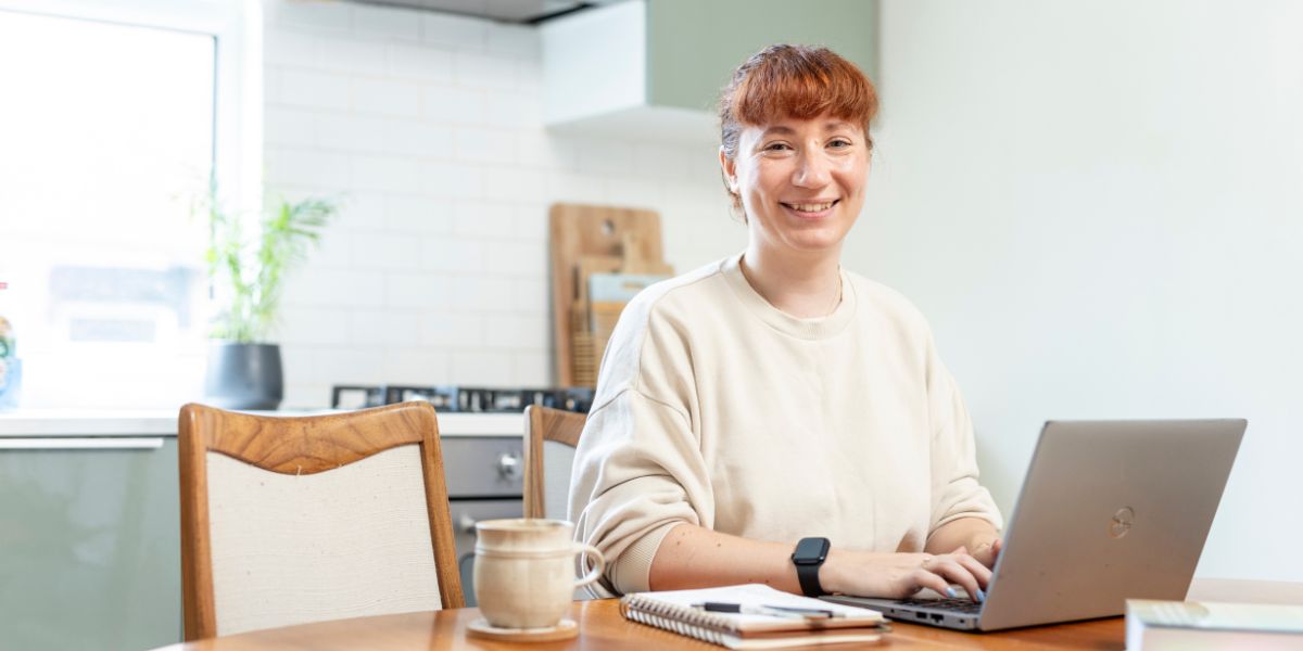 A student working on a laptop at their kitchen table. They are smiling at the camera.