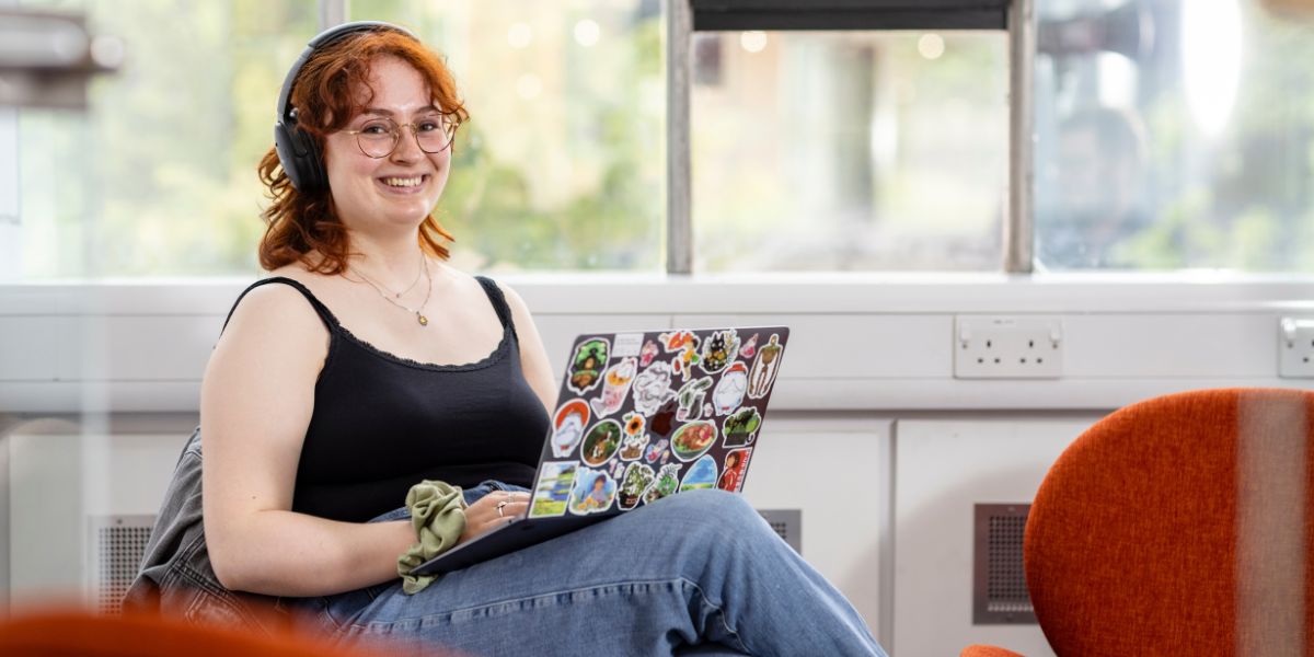 A student sitting in a comfy chair, wearing headphones, with a laptop covered in colourful stickers on their knee.