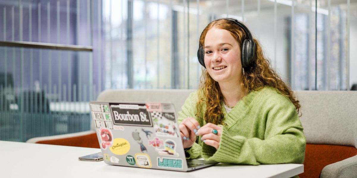 A student, wearing headphones, sits at a table with her laptop. They are smiling.