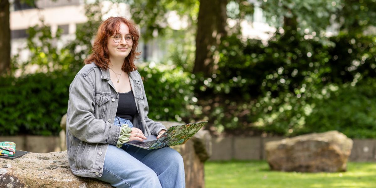 A student sat outside in a tree-lined green space, with a laptop on their knee.