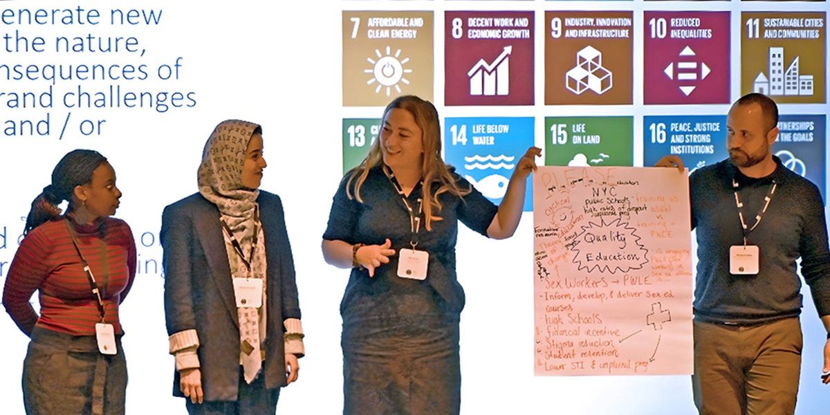 Four people presenting notes written on a large sheet of paper, stood in front of a large screen with a presentation slide on it about the United Nations (UN) Sustainable Development Goals.