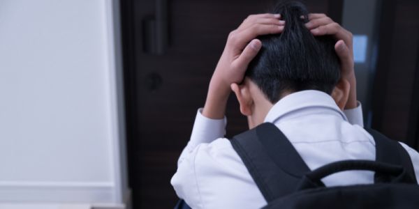 A child with their back to camera, wearing a white shirt and a black backpack. They hold their head in a stressed fashion.