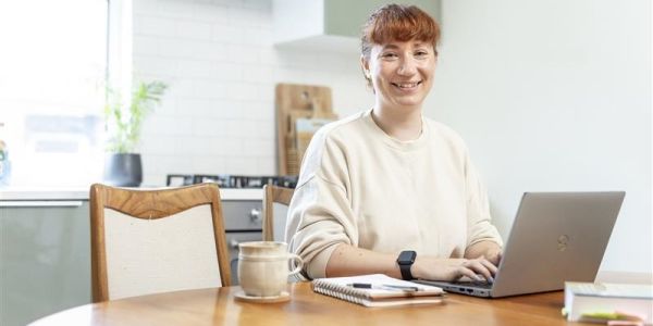 A person sat in a light and bright kitchen working on a laptop at a table, smiling.