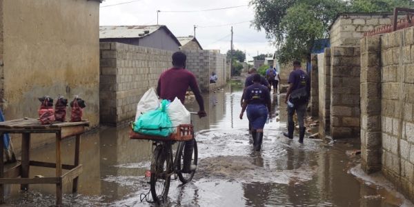 A person cycling through a flooded street with people walking through the flooded street in Africa in the background.