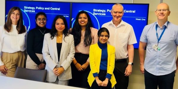 A group of students with a panel of experts from the DWP stood infront of a presentation on a board smiling at the camera.