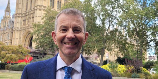 An outdoor image of Professor Mark Mon-Williams, wearing a blue suit and tie, smiling directly into camera. In the background is greenery and the Houses of Parliament.