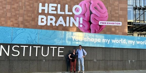 Two people in front of a museum with a graphic of a brain on the wall and the text: Hello Brain