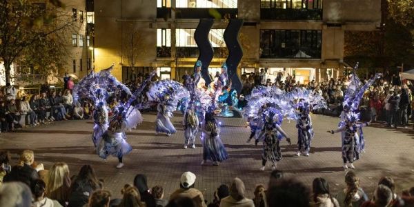 Dancers wearing carnival costumes sway to music in front of the Wavy Bacon statue on the University of Leeds campus.