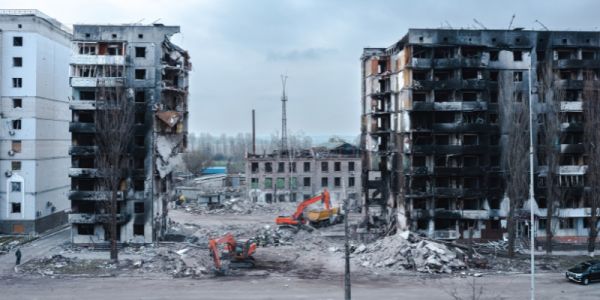 Fire-blackened multi-storey buildings destroyed by war in Ukraine, stand in rubble, beside diggers.