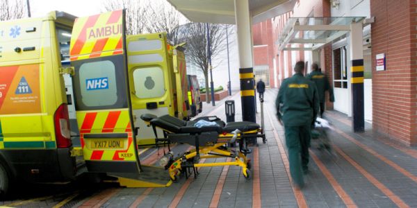 An ambulance parked outside Accident and Emergency (A&E) with its back doors open and a stretcher on wheels nearby, as two paramedics walk towards the A&E entrance.