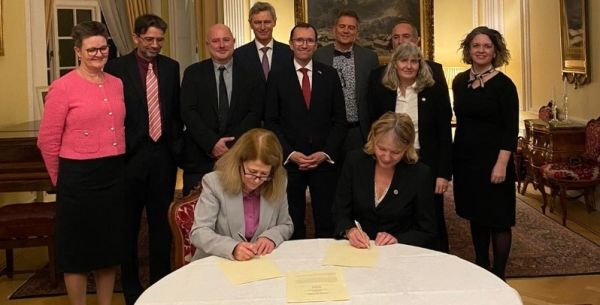 Two people signing a letter of intent with a group of people stood around the table watching them.