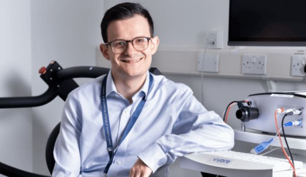 An image of Dr Alexios Dosis smiling and leaning against a desk with research equipment in the background.