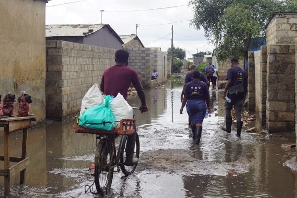 A person cycling through a flooded street with people walking through the flooded street in Africa in the background.