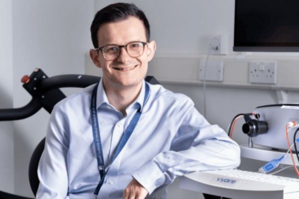 Dr Alexios Dosis smiling and leaning against a desk with research equipment in the background.