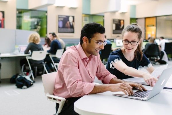 Two smiling students sitting at a desk in a study space using a laptop.