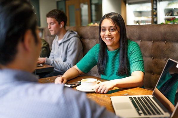 A student enjoying a coffee in the Edit Room cafe. There is a laptop on the desk.