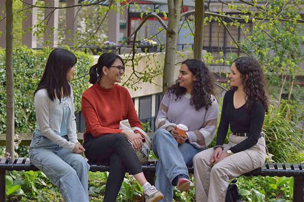 Four students sat on a bench on campus, surrounded by green trees.