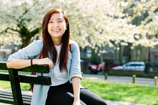 A smiling student sitting on a bench, with blossoming trees and grass behind them.