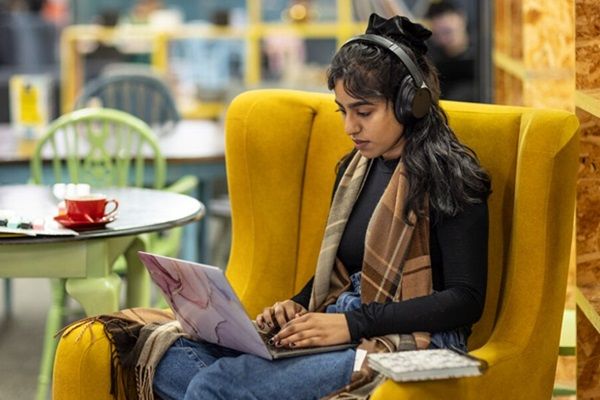 A student sat in a big comfy chair in cafe, wearing headphones and using a laptop.