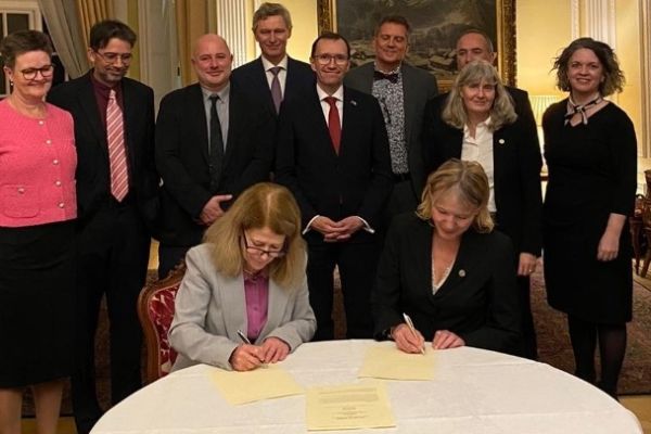 Two people signing a letter of intent with a group of people stood around the table watching them.