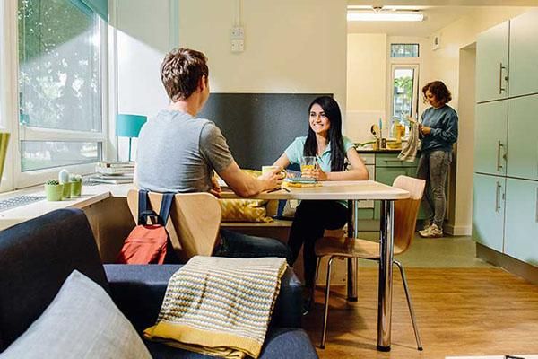 Students in halls of residence. Two are sat at a table, chatting, eating and drinking. Another is washing up in the background.