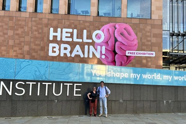 Two people stood in front of a building. Behind them is a graphic of a brain and text that says "Hello Brian" on the building wall.