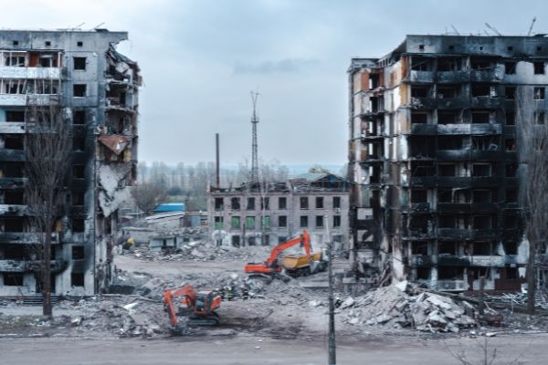 Fire-blackened multi-storey buildings destroyed by war in Ukraine, stand in rubble, beside diggers.