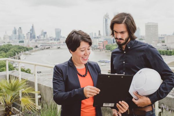 A man and a woman stood on a balcony with a cityscape behind them, looking at a computer tablet.