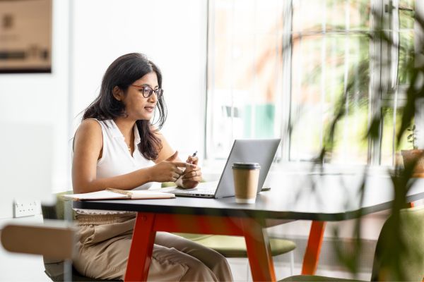 A student sitting at a table in a cafe, taking part in a video call on a laptop.