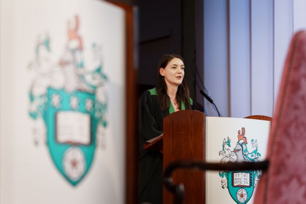 A student in graduation robes speaking at a podium, with the the University of Leeds crest on display on the stage.