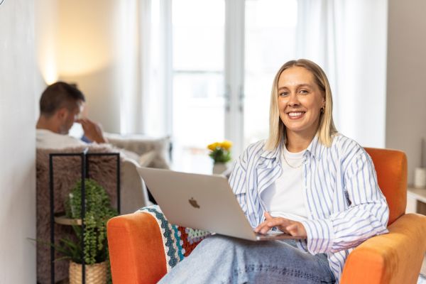 A person sat in an armchair, working on a laptop. They are smiling at the camera.