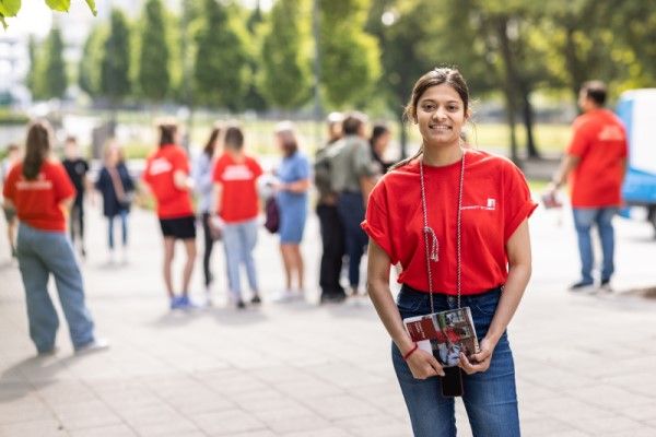 A Leeds student ambassador standing on campus, smiling, holding a brochure, with other student ambassadors and visitors in the background.