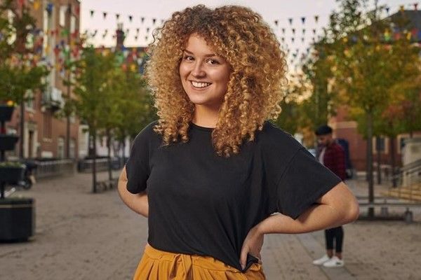 A student standing with their hands on their hips and smiling, in front of Leeds University Union and colourful flags.